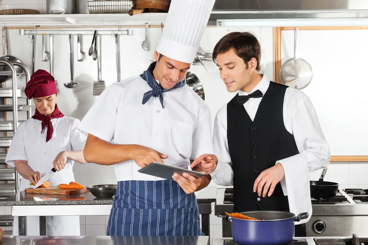 Chef and waiter reading a tablet in a restaurant kitchen while a cook chops carrots